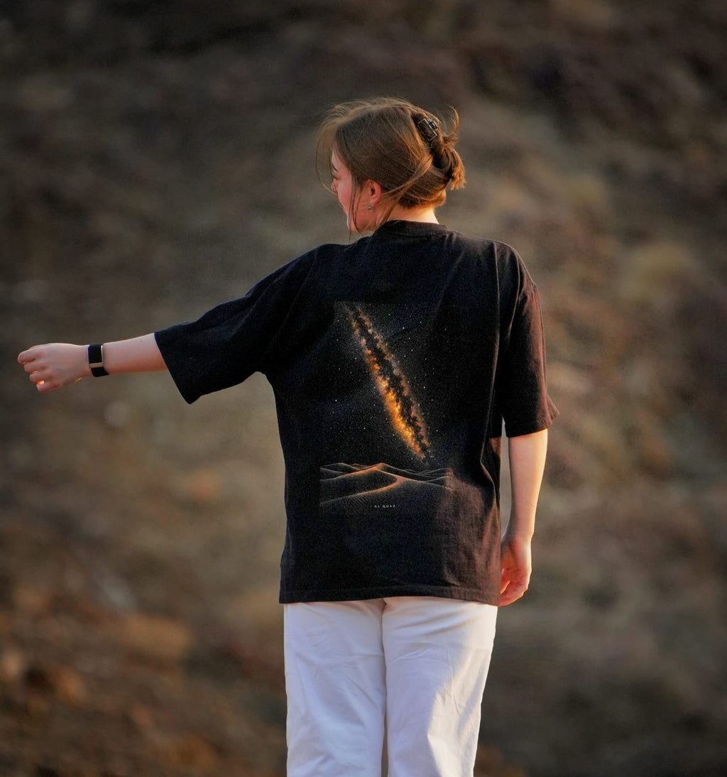 The back of the Al Quaa Galaxy tee in the mountains at dusk, the Milky Way print catching the last light. Female model wears M in the Oversized fit.