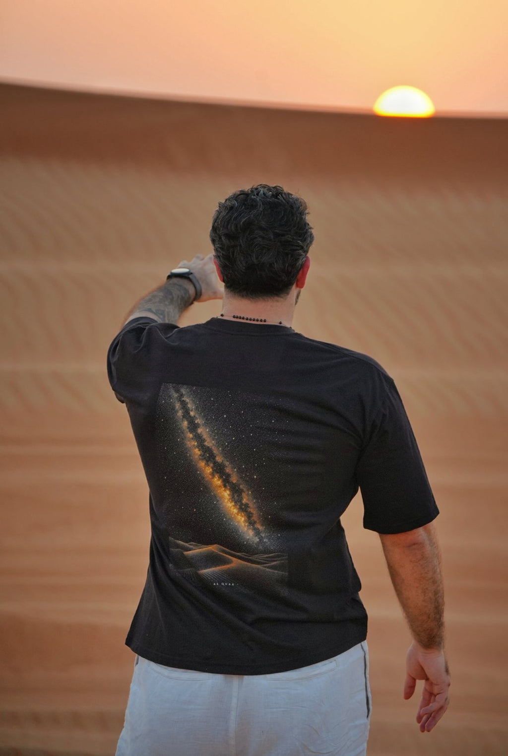 The Al Quaa Galaxy tee worn at sunset — the Milky Way print across the back as the sun drops behind the dunes. Male model is 182 cm, wearing L in the Oversized fit.