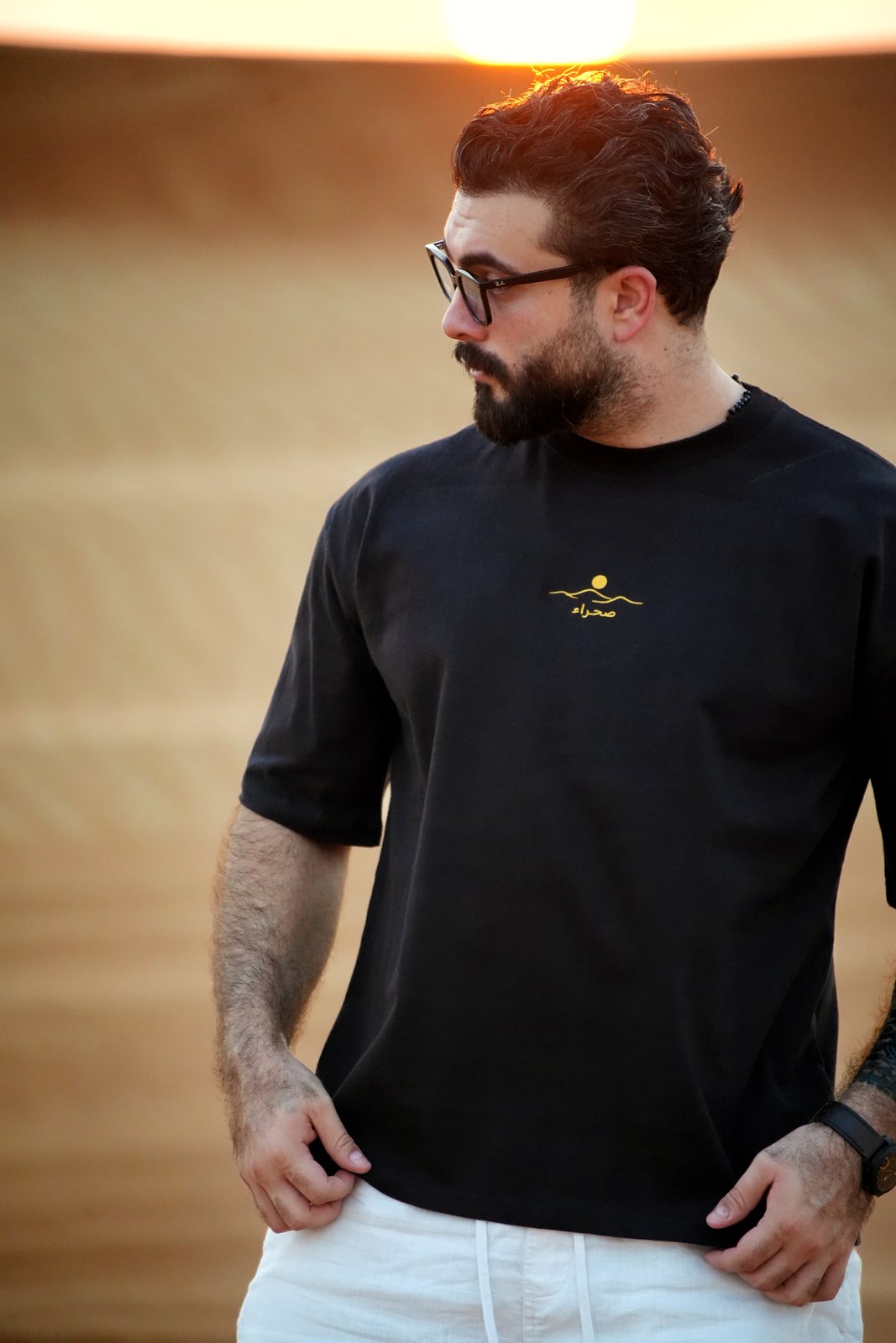 The Al Quaa Galaxy tee worn in the dunes — the gold embroidered sun-over-dunes mark and صحراء on the chest. Male model is 182 cm, wearing L in the Oversized fit.