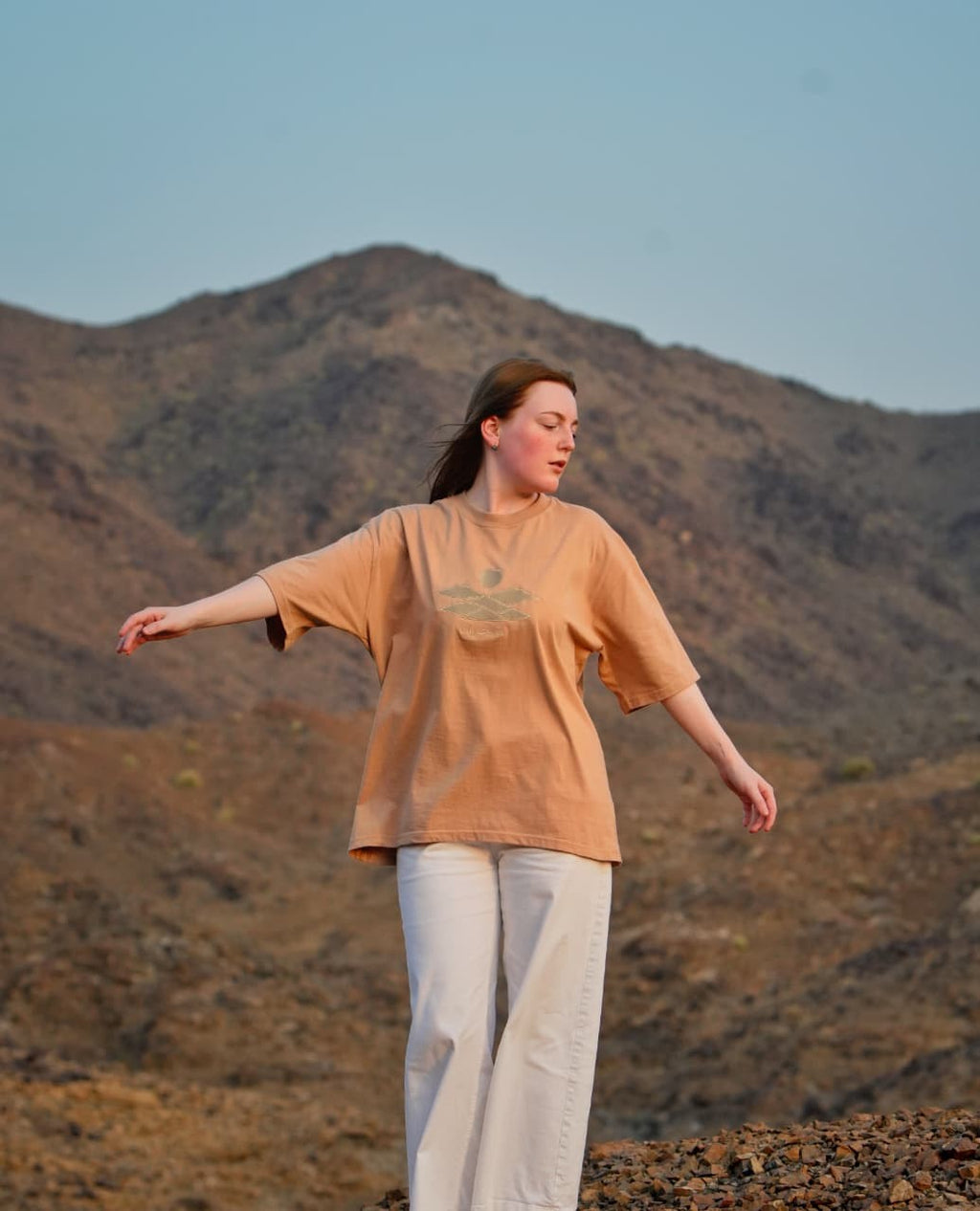 The Empty Quarter tee worn in the mountains, arms out — the tonal embroidery across the chest. Female model wears M in the Oversized fit.