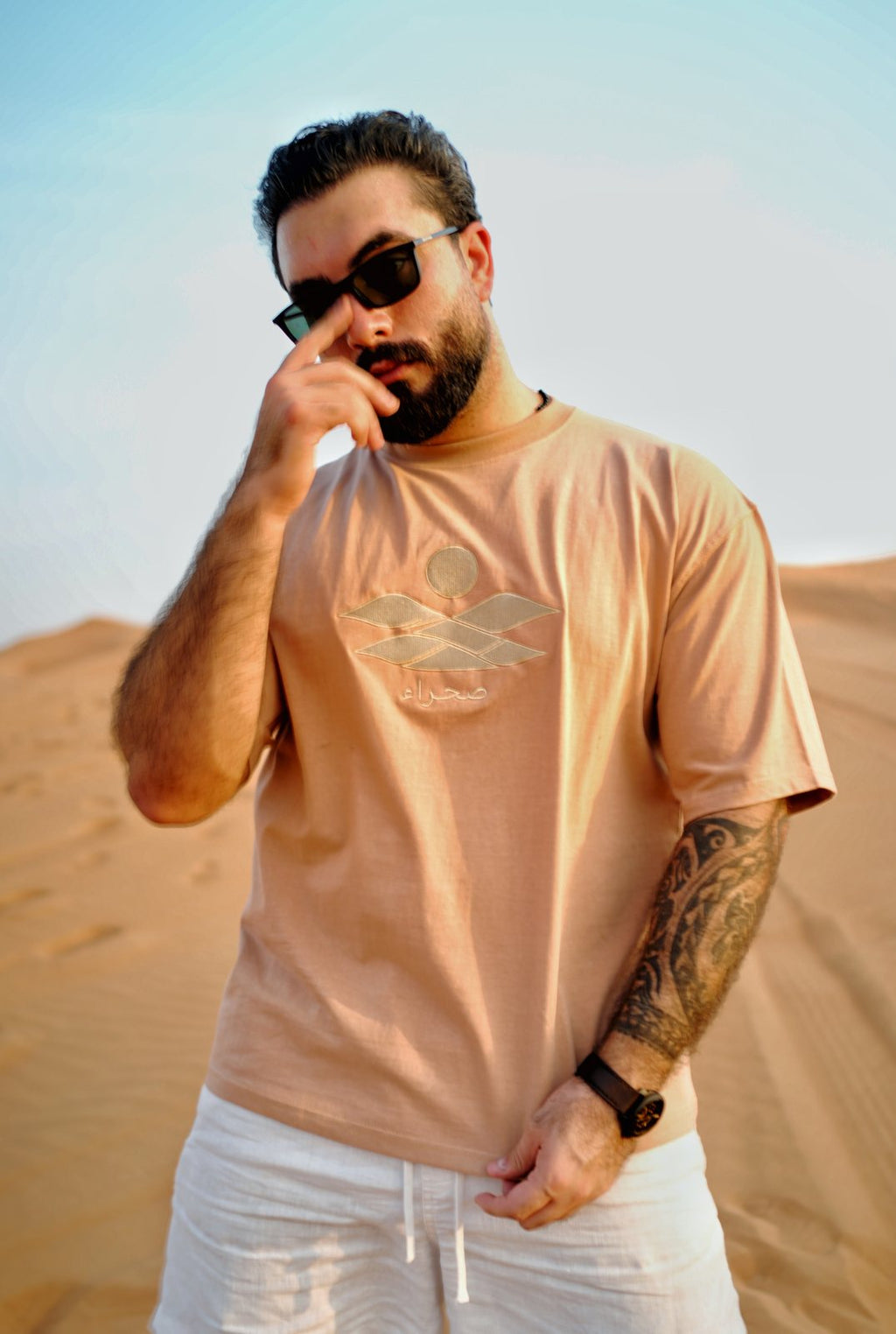 The Empty Quarter tee worn in the dunes — tonal embroidered dunes, sun and صحراء wordmark across the chest. Male model is 182 cm, wearing L in the Oversized fit.