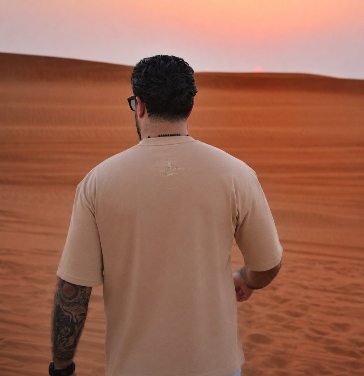 The back of the Empty Quarter tee at sunset, back-neck embroidery visible over the dunes. Male model is 182 cm, wearing L in the Oversized fit.