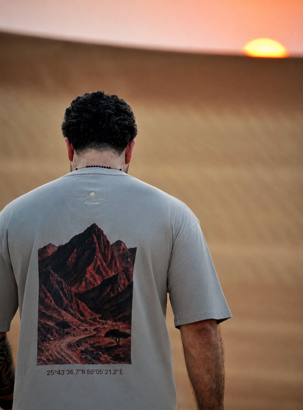 The Hajar Mountains tee worn at sunset — the mountain print and Wadi Naqab coordinates across the back. Male model is 182 cm, wearing L in the Oversized fit.