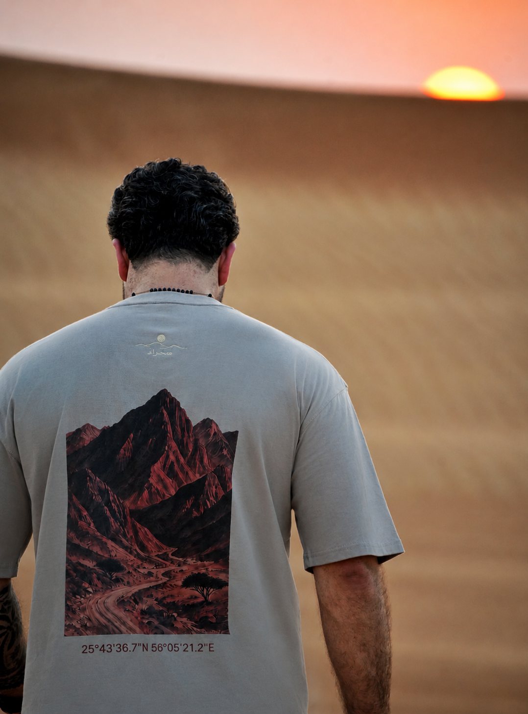 The Hajar Mountains tee worn at sunset — the mountain print and Wadi Naqab coordinates across the back. Male model is 182 cm, wearing L in the Oversized fit.