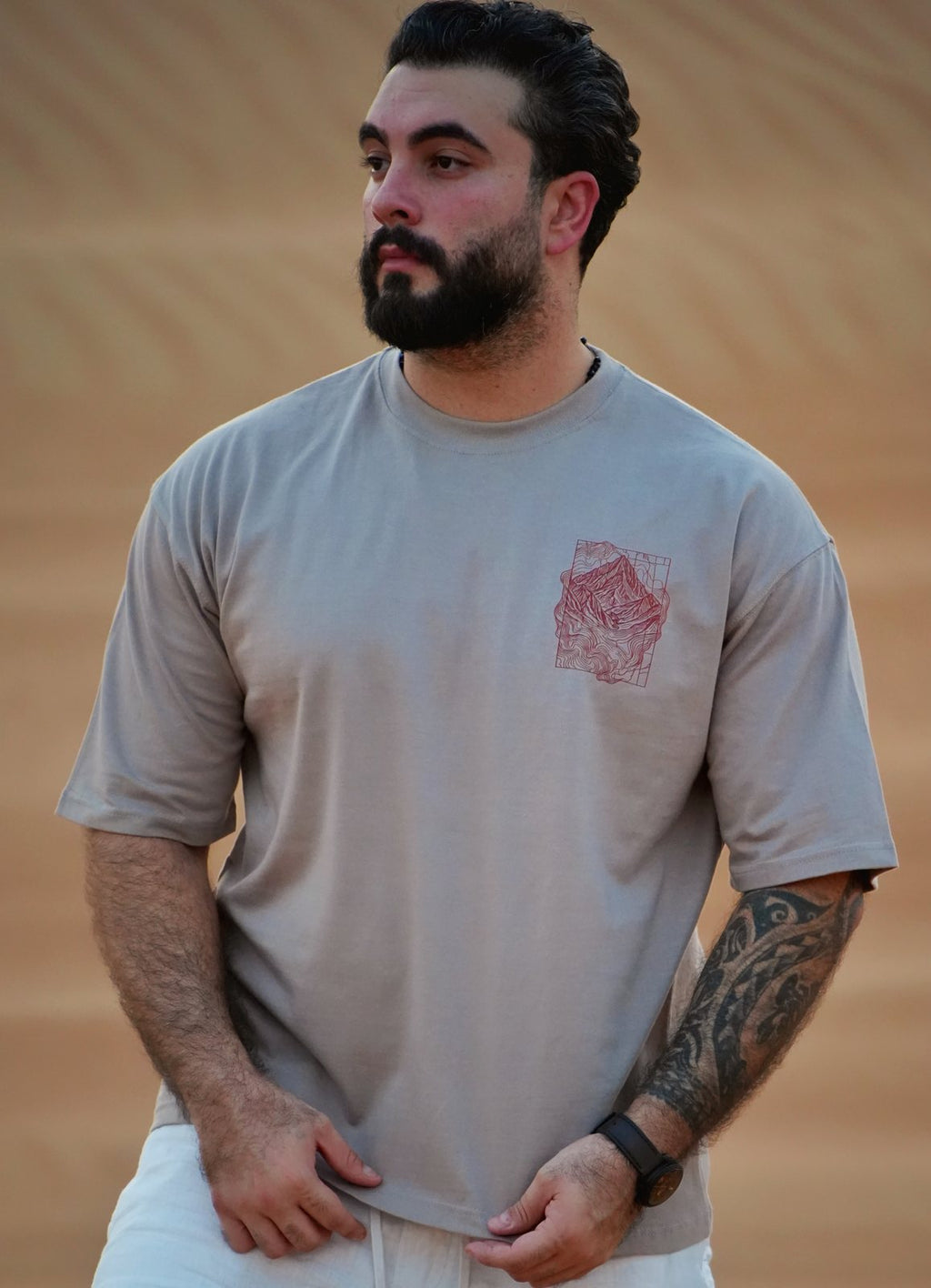 The Hajar Mountains tee worn in the dunes — the contour-line mountain print on the chest. Male model is 182 cm, wearing L in the Oversized fit.