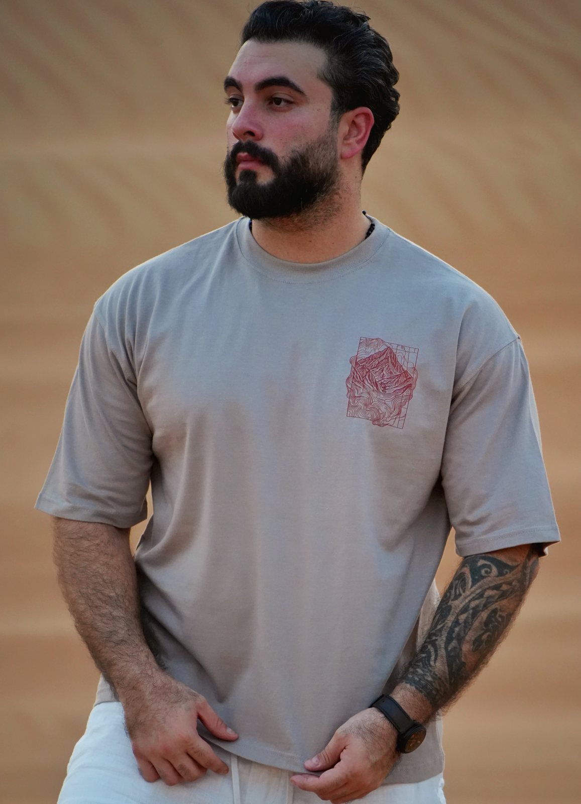 The Hajar Mountains tee worn in the dunes — the contour-line mountain print on the chest. Male model is 182 cm, wearing L in the Oversized fit.