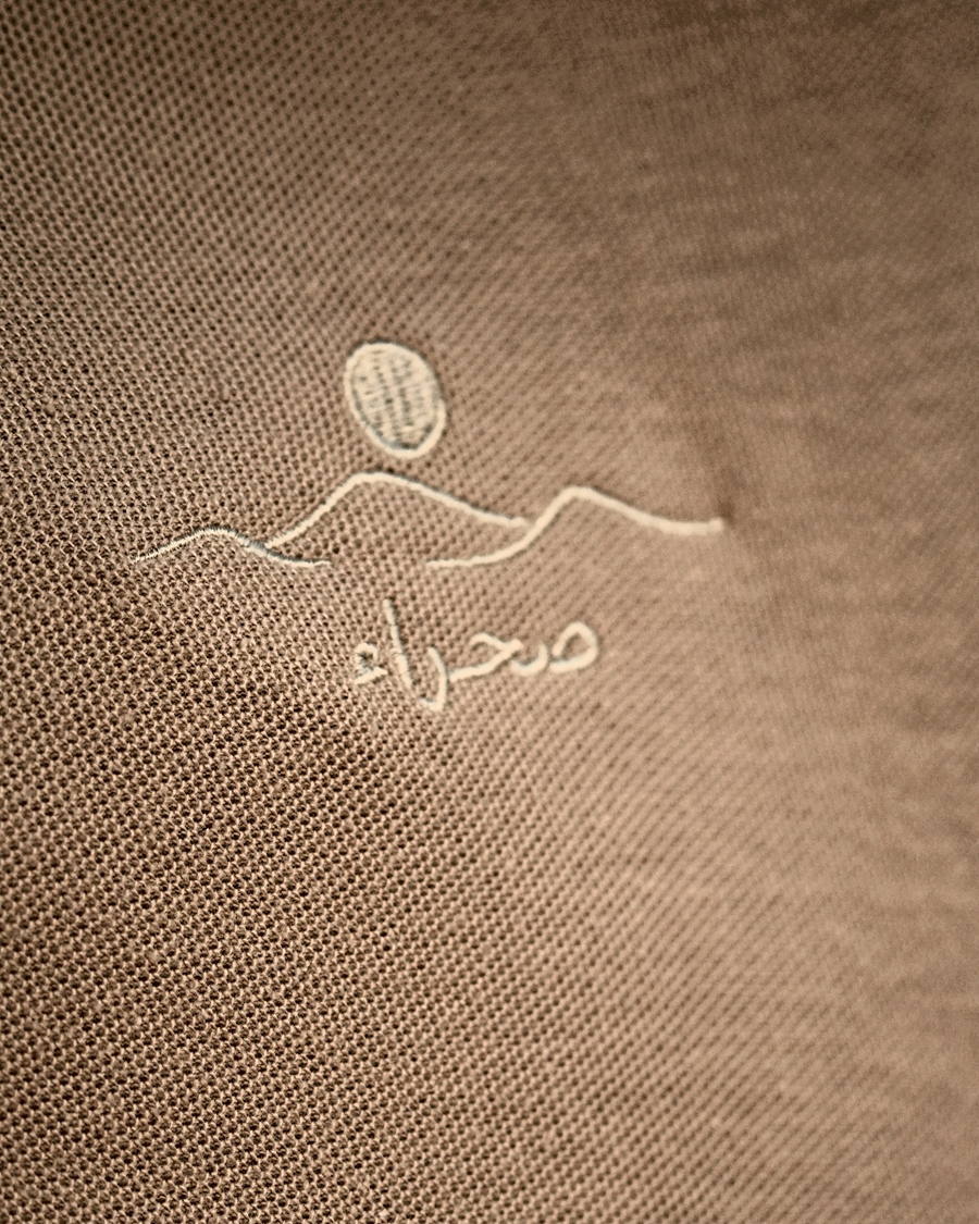 Close-up of the embroidery on The Sahra Polo — the dune line, sun and صحراء stitched in cream thread on sand piqué