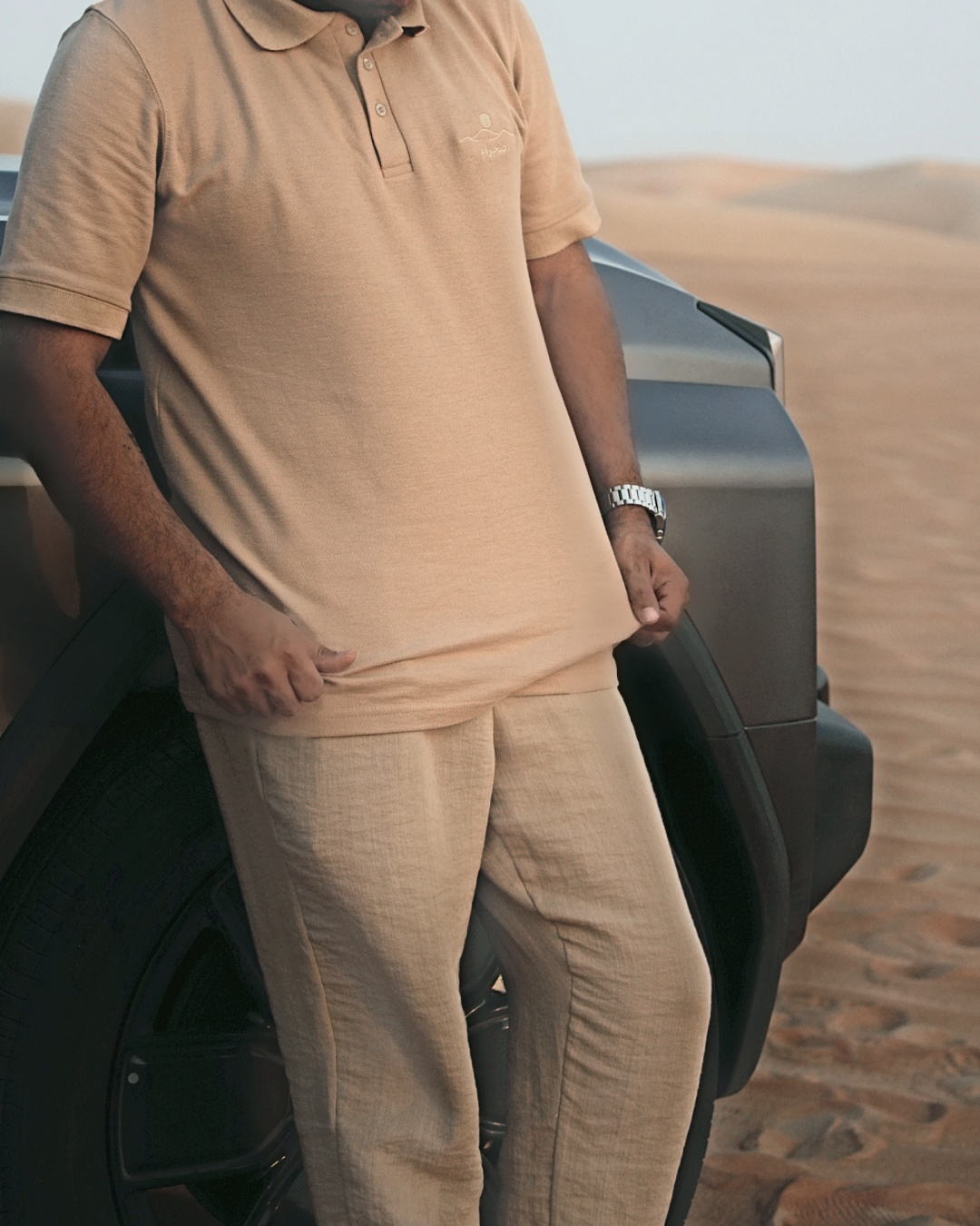 The Sahra Polo in sand, worn against a 4x4 in the dunes — 240gsm piqué cotton, three-button placket, embroidered mark on the chest